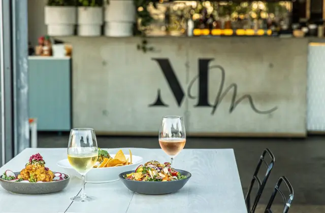A white outdoor table set with a glass of white wine, a glass of rosé and three share dishes — fried cauliflower with pomegranate, a bowl of corn chips and a leafy salad — in front of a concrete bar wall painted with the Mary Mae's monogram, with potted herbs on the counter behind.