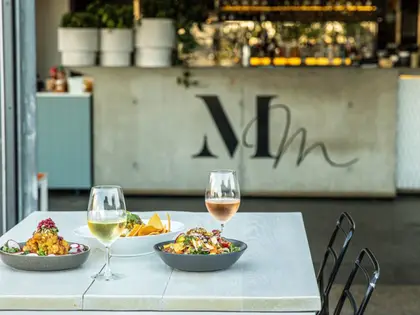 A white outdoor table set with a glass of white wine, a glass of rosé and three share dishes — fried cauliflower with pomegranate, a bowl of corn chips and a leafy salad — in front of a concrete bar wall painted with the Mary Mae's monogram, with potted herbs on the counter behind.