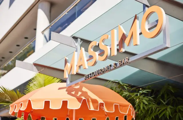 Looking up from the footpath at cut-out cream lettering reading MASSIMO RESTAURANT & BAR, hung from a steel frame under a glass awning in bright sun, with the scalloped edge of an orange umbrella and palm and bamboo foliage below.