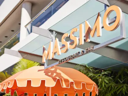 Looking up from the footpath at cut-out cream lettering reading MASSIMO RESTAURANT & BAR, hung from a steel frame under a glass awning in bright sun, with the scalloped edge of an orange umbrella and palm and bamboo foliage below.
