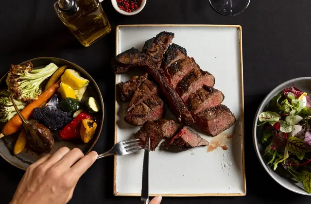 Overhead view of a black-clothed table: a charred T-bone steak carved into thick pink-centred slices on a rectangular plate, a hand holding a fork and steak knife at the edge, a bowl of roasted vegetables — carrots, cauliflower, courgette, baby capsicum and eggplant — to one side, a leaf salad to the other, and a bottle of olive oil, a dish of pink and black peppercorns and an empty wine glass at the top of the frame.