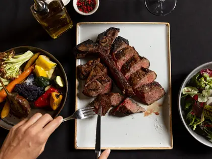 Overhead view of a black-clothed table: a charred T-bone steak carved into thick pink-centred slices on a rectangular plate, a hand holding a fork and steak knife at the edge, a bowl of roasted vegetables — carrots, cauliflower, courgette, baby capsicum and eggplant — to one side, a leaf salad to the other, and a bottle of olive oil, a dish of pink and black peppercorns and an empty wine glass at the top of the frame.