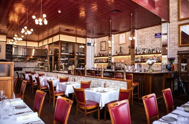A dining room of white-clothed tables and red leather bentwood chairs on dark timber floors, under a pressed-metal ceiling painted deep red with brass sputnik pendant lights. A whitewashed brick bar with a marble top runs down the middle, backed by lit shelves of spirit bottles and glass-fronted wine cabinets, with an open kitchen pass at the far end and a blue French street sign on the wall.