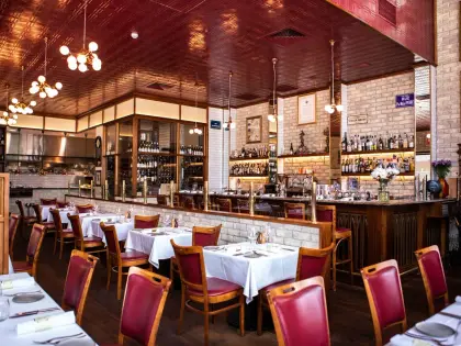 A dining room of white-clothed tables and red leather bentwood chairs on dark timber floors, under a pressed-metal ceiling painted deep red with brass sputnik pendant lights. A whitewashed brick bar with a marble top runs down the middle, backed by lit shelves of spirit bottles and glass-fronted wine cabinets, with an open kitchen pass at the far end and a blue French street sign on the wall.