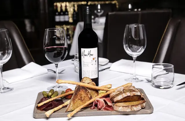 A charcuterie board on a white-clothed restaurant table holding thin slices of cured meat, prosciutto, cornichons, grissini and toasted and rustic bread, with a bottle of Moo Moo "Dancing Cow" Shiraz and a poured glass of red wine behind it.
