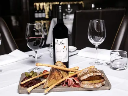 A charcuterie board on a white-clothed restaurant table holding thin slices of cured meat, prosciutto, cornichons, grissini and toasted and rustic bread, with a bottle of Moo Moo "Dancing Cow" Shiraz and a poured glass of red wine behind it.