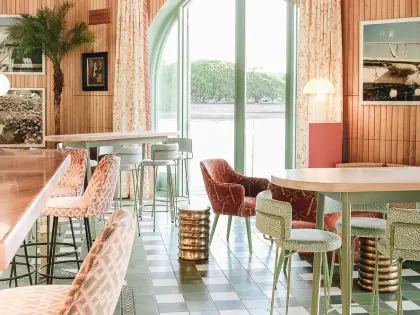 A riverside room in pinks and greens: timber-panelled walls hung with framed photographs, patterned pink and mint upholstered chairs and bar stools around round timber tables, brass drum-shaped side tables, a green and white chequerboard tiled floor, and tall arched glass doors standing open to the water and the trees on the far bank.