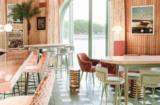 A riverside room in pinks and greens: timber-panelled walls hung with framed photographs, patterned pink and mint upholstered chairs and bar stools around round timber tables, brass drum-shaped side tables, a green and white chequerboard tiled floor, and tall arched glass doors standing open to the water and the trees on the far bank.