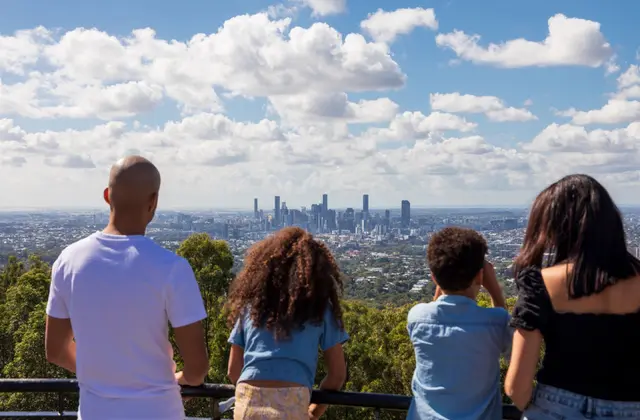 Four people seen from behind at a black metal railing on a hilltop, looking out across bushland treetops and suburban rooftops to a cluster of city towers on the horizon; one raises a phone to take a photograph, under a blue sky banked with white cloud.