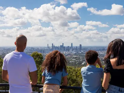 Four people seen from behind at a black metal railing on a hilltop, looking out across bushland treetops and suburban rooftops to a cluster of city towers on the horizon; one raises a phone to take a photograph, under a blue sky banked with white cloud.