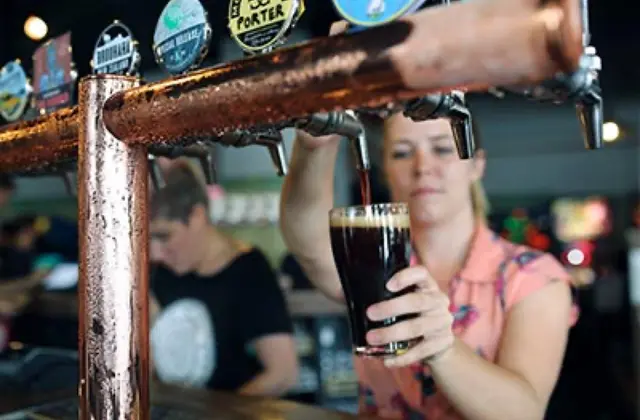 Close-up of a copper tap bank on the bar, beer badges mounted above it, as a bartender in a pink patterned shirt pulls a glass of dark beer. Another staff member works further along the bar in soft focus.