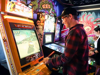 A man in a red flannel shirt and dark cap plays a yellow-sided arcade cabinet showing a driving game, gripping the controls in profile. Pinball machines and a wall of psychedelic mural art in pinks and greens fill the room behind him.