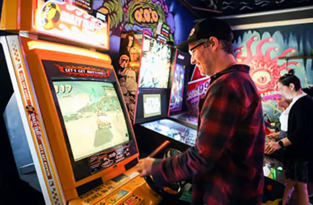 A man in a red flannel shirt and dark cap plays a yellow-sided arcade cabinet showing a driving game, gripping the controls in profile. Pinball machines and a wall of psychedelic mural art in pinks and greens fill the room behind him.