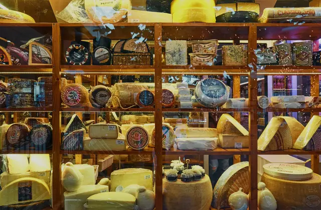 A lit, glass-fronted timber shelving cabinet packed with cheese — whole wheels, cut wedges, blue-veined slabs, cloth- and wax-wrapped rounds, each with a small paper label — with cured meats on the top shelf and parcels of mozzarella and pale rounds on the bottom; the street outside is reflected in the glass.