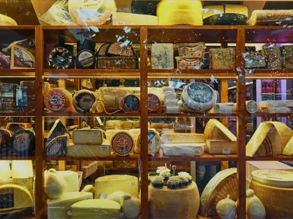 A lit, glass-fronted timber shelving cabinet packed with cheese — whole wheels, cut wedges, blue-veined slabs, cloth- and wax-wrapped rounds, each with a small paper label — with cured meats on the top shelf and parcels of mozzarella and pale rounds on the bottom; the street outside is reflected in the glass.
