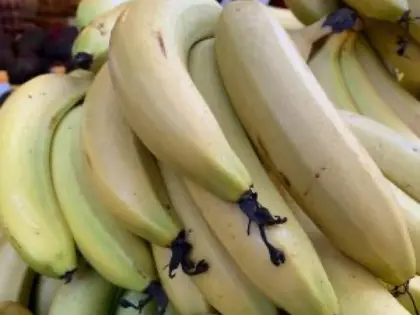 A close-up pile of pale green-yellow bananas still in hands, stacked on a market stall with other fruit and a stallholder blurred behind.