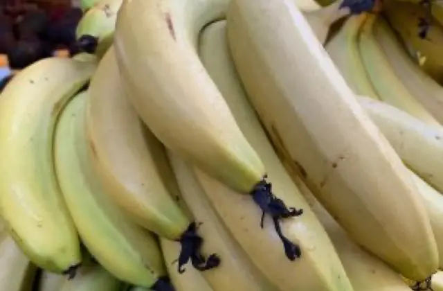 A close-up pile of pale green-yellow bananas still in hands, stacked on a market stall with other fruit and a stallholder blurred behind.
