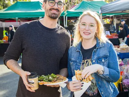 A man holding a takeaway coffee cup and a plate of topped flatbread stands beside a woman in a denim jacket holding a wedge of pastry in a paper sleeve, in front of green market marquees.