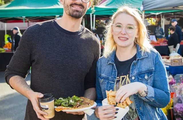 A man holding a takeaway coffee cup and a plate of topped flatbread stands beside a woman in a denim jacket holding a wedge of pastry in a paper sleeve, in front of green market marquees.