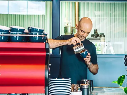 A man in a black T-shirt pouring steamed milk from a steel jug into a dark blue cup, standing behind a pale marble counter. A red espresso machine topped with rows of blue cups stands to his left, a tall stack of blue saucers and a pot of spoons in front of him, and behind him green ribbed glazing and a coffee roaster fill the room.
