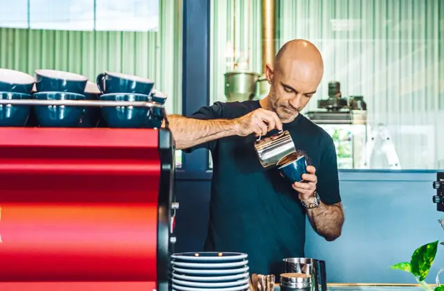A man in a black T-shirt pouring steamed milk from a steel jug into a dark blue cup, standing behind a pale marble counter. A red espresso machine topped with rows of blue cups stands to his left, a tall stack of blue saucers and a pot of spoons in front of him, and behind him green ribbed glazing and a coffee roaster fill the room.