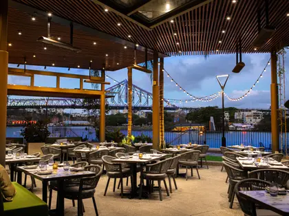 A covered outdoor terrace at dusk with a slatted timber ceiling, downlights, overhead gas heaters and strings of festoon lights, filled with grey woven rattan chairs at set tables, looking across the river to the Story Bridge.