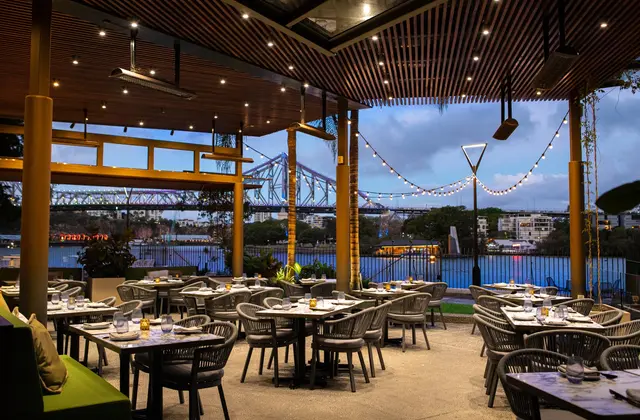 A covered outdoor terrace at dusk with a slatted timber ceiling, downlights, overhead gas heaters and strings of festoon lights, filled with grey woven rattan chairs at set tables, looking across the river to the Story Bridge.
