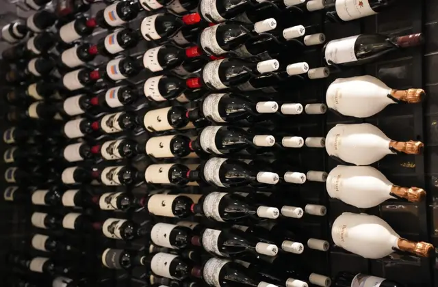 A dark wall of wine racks holding rows of bottles lying on their sides, mostly red wine with dark glass and pale labels, with several large Ruinart champagne bottles in white sleeves and gold foil tops standing out on the right.