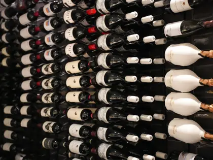 A dark wall of wine racks holding rows of bottles lying on their sides, mostly red wine with dark glass and pale labels, with several large Ruinart champagne bottles in white sleeves and gold foil tops standing out on the right.
