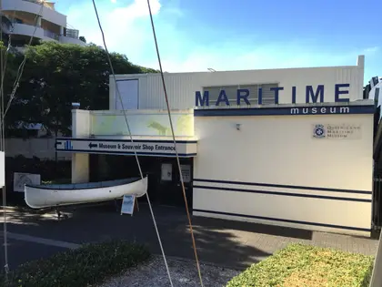 The museum's cream entrance building with large blue MARITIME museum lettering along the top, a white timber lifeboat on a cradle beside the doorway and a sign pointing to the museum and souvenir shop.