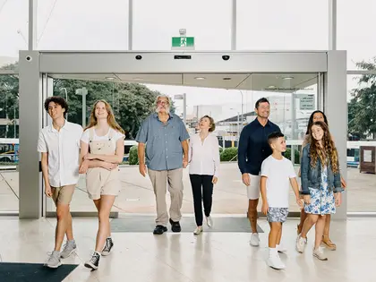 Eight people of mixed ages — teenagers, adults and two young children — walking out through the museum's wide glass sliding doors onto a pale tiled forecourt, with "Welcome to Queensland Museum" showing in reverse on the glass behind them.