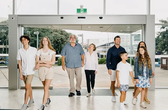 Eight people of mixed ages — teenagers, adults and two young children — walking out through the museum's wide glass sliding doors onto a pale tiled forecourt, with "Welcome to Queensland Museum" showing in reverse on the glass behind them.