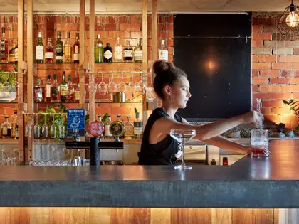 A bartender in a black singlet stirring a drink in a tall glass at a long concrete-topped bar, an empty coupe glass beside her. Behind her are beer taps with brewery badges, timber shelves of spirit bottles, upturned stemware in racks and trailing pot plants against a warmly lit exposed red brick wall, with a blackboard and a caged filament bulb at the right.