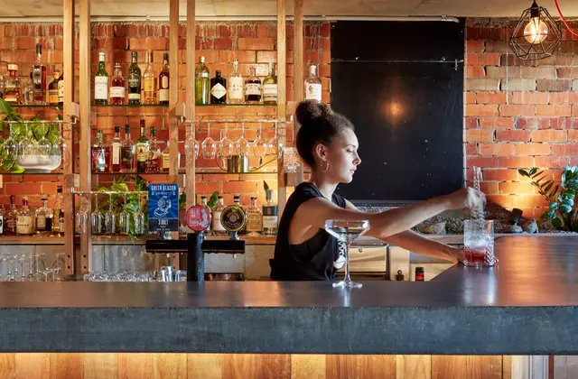 A bartender in a black singlet stirring a drink in a tall glass at a long concrete-topped bar, an empty coupe glass beside her. Behind her are beer taps with brewery badges, timber shelves of spirit bottles, upturned stemware in racks and trailing pot plants against a warmly lit exposed red brick wall, with a blackboard and a caged filament bulb at the right.