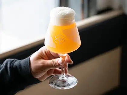 A hand in a black sleeve holding up a stemmed tulip glass of hazy orange beer with a thick white head, etched with a small diamond-shaped mark, in front of a bright window and a black banquette.