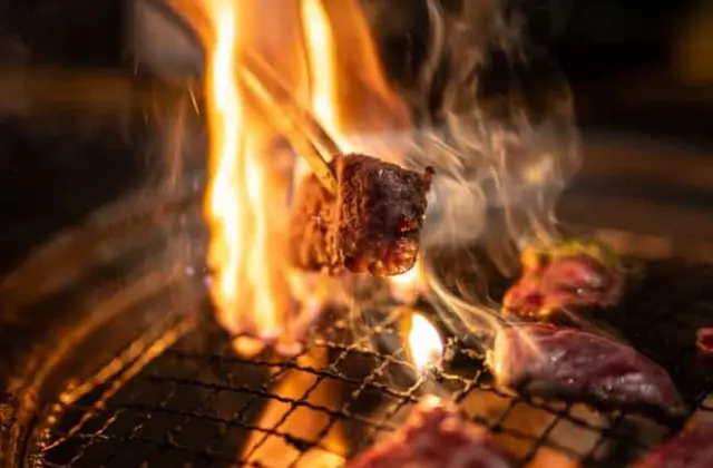 Close-up of a round tabletop charcoal grill in a dark room: flame licks up through the wire mesh and chopsticks lift a caramelised cube of marbled beef into the smoke, with more raw sliced beef laid across the grate.
