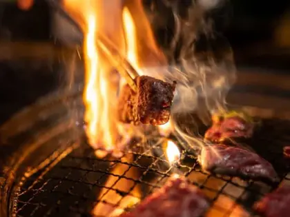 Close-up of a round tabletop charcoal grill in a dark room: flame licks up through the wire mesh and chopsticks lift a caramelised cube of marbled beef into the smoke, with more raw sliced beef laid across the grate.