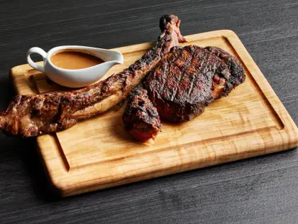 A grilled bone-in steak with dark char lines, its meat carved off the long rib bone and laid beside it, on a grooved wooden board with a small white jug of brown sauce, against a dark grey timber surface.