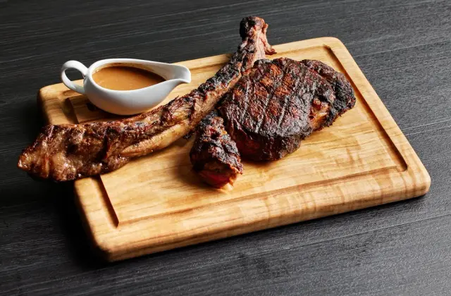 A grilled bone-in steak with dark char lines, its meat carved off the long rib bone and laid beside it, on a grooved wooden board with a small white jug of brown sauce, against a dark grey timber surface.