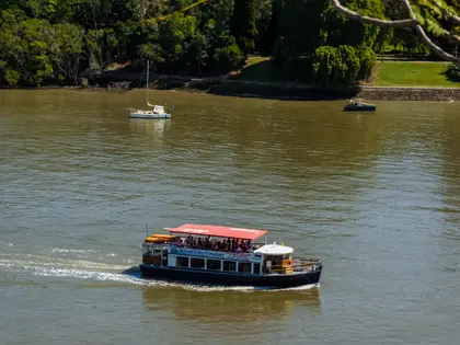 A small blue-hulled cruise boat with a red canopy motoring up the brown river, 'River City Cruises' lettered along its side and passengers seated on the open upper deck, with a moored yacht and a green wooded bank behind.