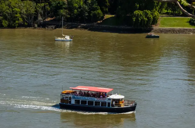 A small blue-hulled cruise boat with a red canopy motoring up the brown river, 'River City Cruises' lettered along its side and passengers seated on the open upper deck, with a moored yacht and a green wooded bank behind.