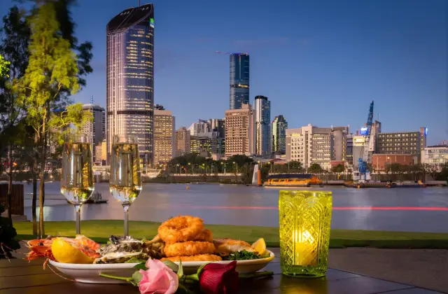 An outdoor table at dusk holding a platter of whole prawns, oysters on the shell, crumbed calamari rings, grilled fish, lemon wedges and greens, with two glasses of sparkling wine, a lit green cut-glass candle holder and a pink and a red rose laid on the table. Beyond is a lawn, the river with a ferry at a terminal, and the lit towers of the city under a deep blue sky.