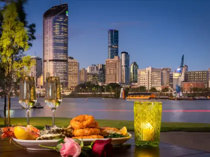 An outdoor table at dusk holding a platter of whole prawns, oysters on the shell, crumbed calamari rings, grilled fish, lemon wedges and greens, with two glasses of sparkling wine, a lit green cut-glass candle holder and a pink and a red rose laid on the table. Beyond is a lawn, the river with a ferry at a terminal, and the lit towers of the city under a deep blue sky.