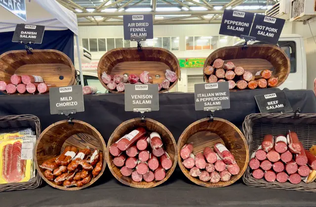 A market stall counter lined with wooden barrels and wicker baskets of cured sausage — salami, chorizo, prosciutto — each with a black label naming the variety, under the market shed's roof.