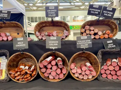 A market stall counter lined with wooden barrels and wicker baskets of cured sausage — salami, chorizo, prosciutto — each with a black label naming the variety, under the market shed's roof.
