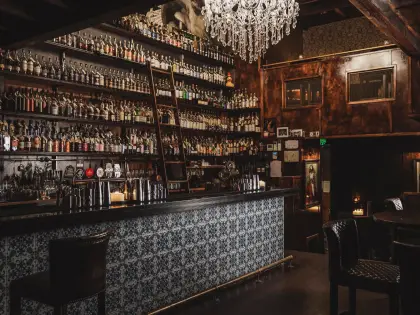 The dimly lit back bar: shelves of spirit bottles stacked to the ceiling with a wooden rolling ladder leaning against them, a crystal chandelier and a marble bust above, and a bar front tiled in blue-and-white patterned tiles with leather stools alongside.