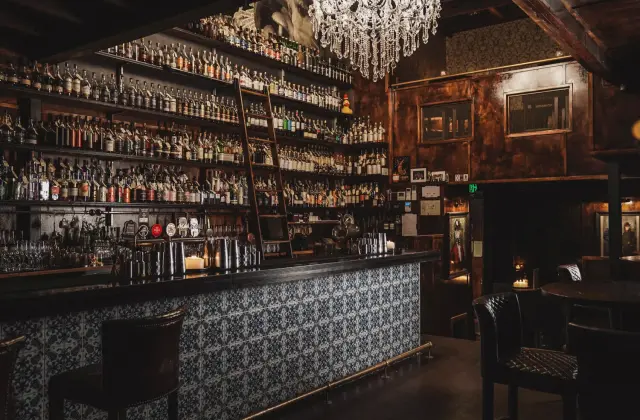 The dimly lit back bar: shelves of spirit bottles stacked to the ceiling with a wooden rolling ladder leaning against them, a crystal chandelier and a marble bust above, and a bar front tiled in blue-and-white patterned tiles with leather stools alongside.
