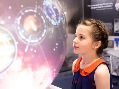 A girl with braided hair, in a navy dress with an orange collar, looking up at a large illuminated display of planets ringed by orbital diagrams against a pink starfield; dark exhibition panels behind her carry photographs of astronauts, a rocket launch and the lunar surface.