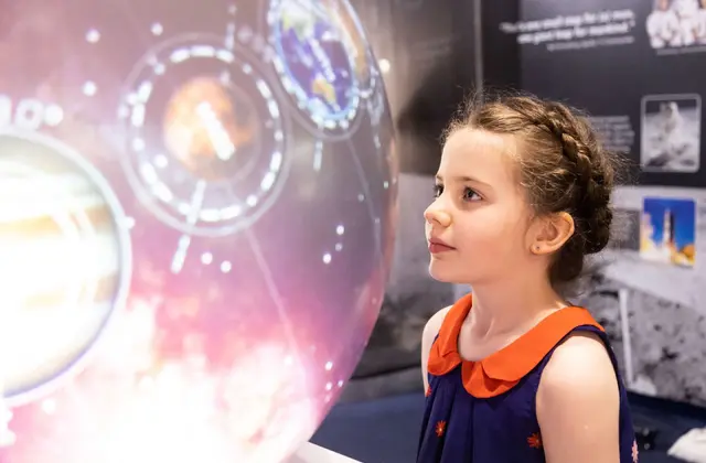 A girl with braided hair, in a navy dress with an orange collar, looking up at a large illuminated display of planets ringed by orbital diagrams against a pink starfield; dark exhibition panels behind her carry photographs of astronauts, a rocket launch and the lunar surface.