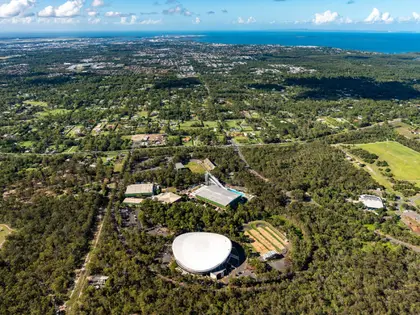 Aerial view of a large white domed velodrome ringed by car parks, with green-roofed aquatic and indoor sports buildings, a diving tower and a dirt BMX track alongside, all set in dense bushland; suburban houses, a golf course and the bay stretch to the horizon under scattered cloud.
